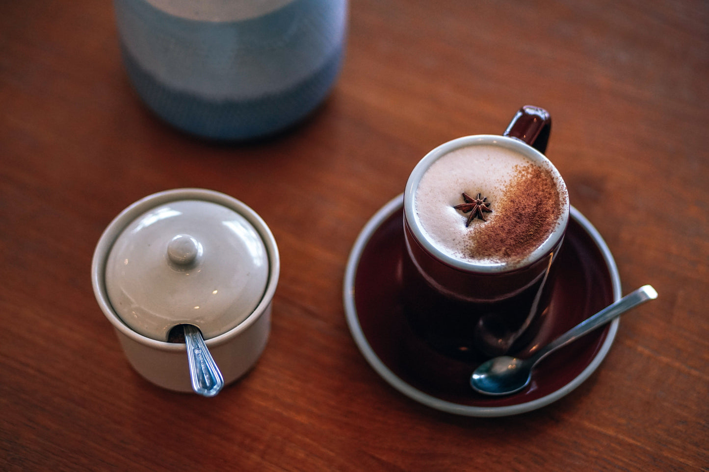 A close-up of a cappuccino in a dark ceramic cup on a matching saucer, topped with foam and a star anise, alongside a small lidded sugar bowl on a wooden table.