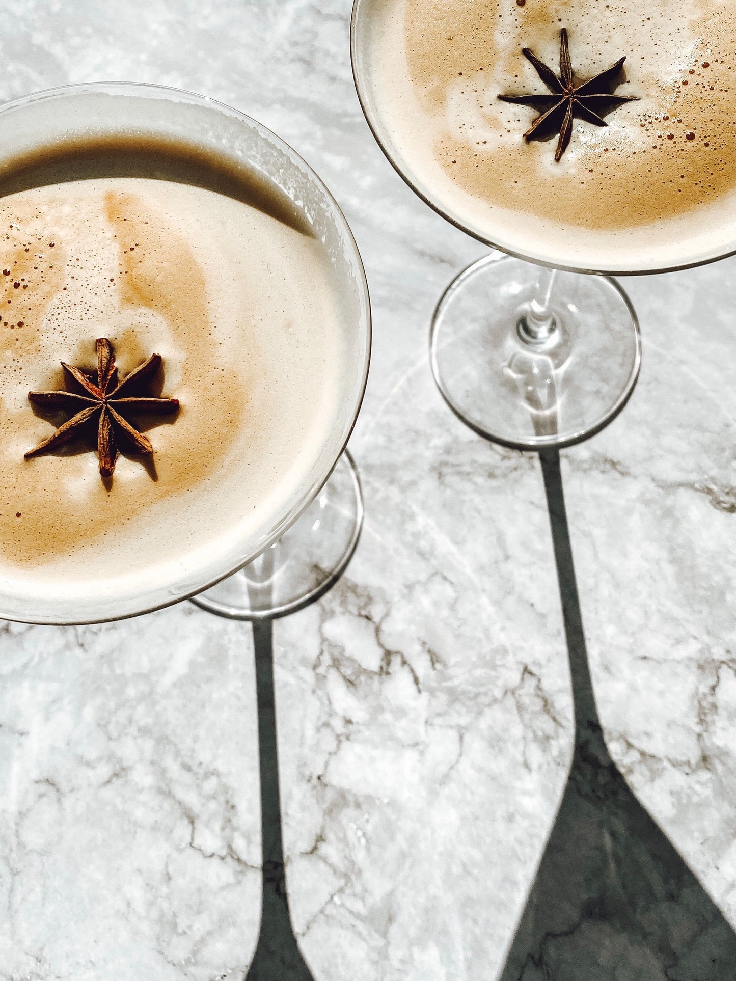 Two stemmed cocktail glasses filled with a light brown beverage topped with foam and a star anise garnish, set on a white marble surface with sharp shadows.
