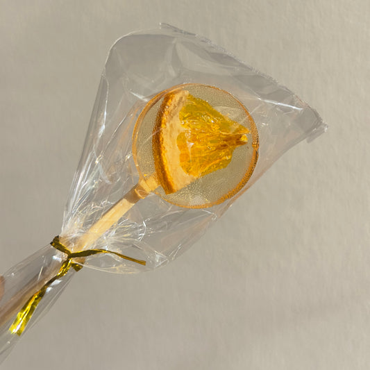 Lollipop with orange slice on a clear stick, wrapped in cellophane against a neutral background