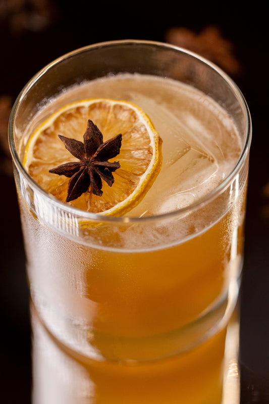 A close-up of a clear glass filled with an amber-colored cocktail, topped with ice, a lemon slice, and a star anise garnish against a dark background.